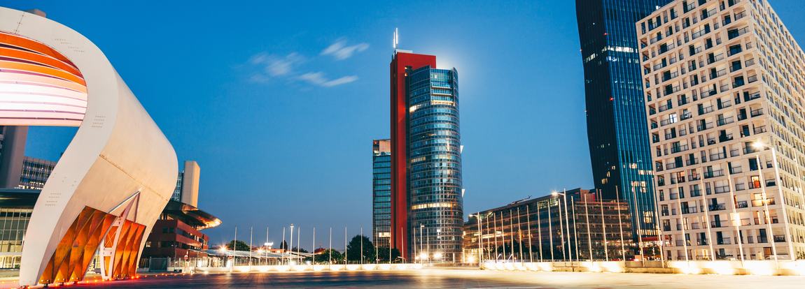 Bildbeschreibung: Moderne Architektur bei Nacht. Im Vordergrund steht ein eleganter, geschwungener Bau mit metallischen Elementen. Im Hintergrund ragen Hochh&auml;user mit gl&auml;sernen Fassaden in den Himmel, w&auml;hrend die Umgebung sanft beleuchtet ist. Der Himmel ist klar und blau.