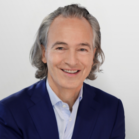 Aurel Schoeller - President A man with medium-length, wavy gray hair smiles warmly. He wears a dark blue blazer over a light blue shirt. The background is light and simple, emphasizing his friendly expression.