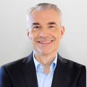 Ingmar Köhler - Vice President Production Ein lächelnder Mann mit grauen Haaren trägt einen schwarzen Anzug und ein hellblaues Hemd. Der Hintergrund ist neutral und hell.