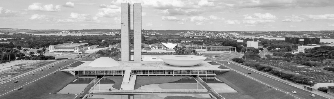Das Bild zeigt das Nationalkongressgeb&auml;ude in Bras&iacute;lia, Brasilien. Es besteht aus zwei T&uuml;rmen und einem gro&szlig;en, runden Geb&auml;ude im Vordergrund. Die Architektur ist modern und prominent, umgeben von einer weiten, gr&uuml;nen Landschaft und einem Himmel mit Wolken.