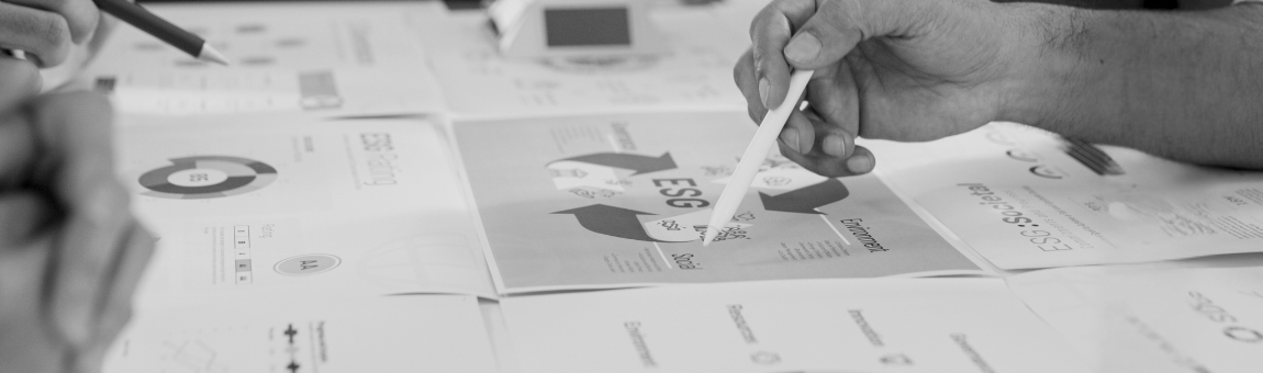 Hände, die an einem Tisch arbeiten, zeigen verschiedene Papierdokumente mit Diagrammen und Texten. Ein handgehaltenes Schreibgerät deutet auf ein Dokument mit dem Thema "ESG" (Umwelt, Soziales, Unternehmensführung) hin.